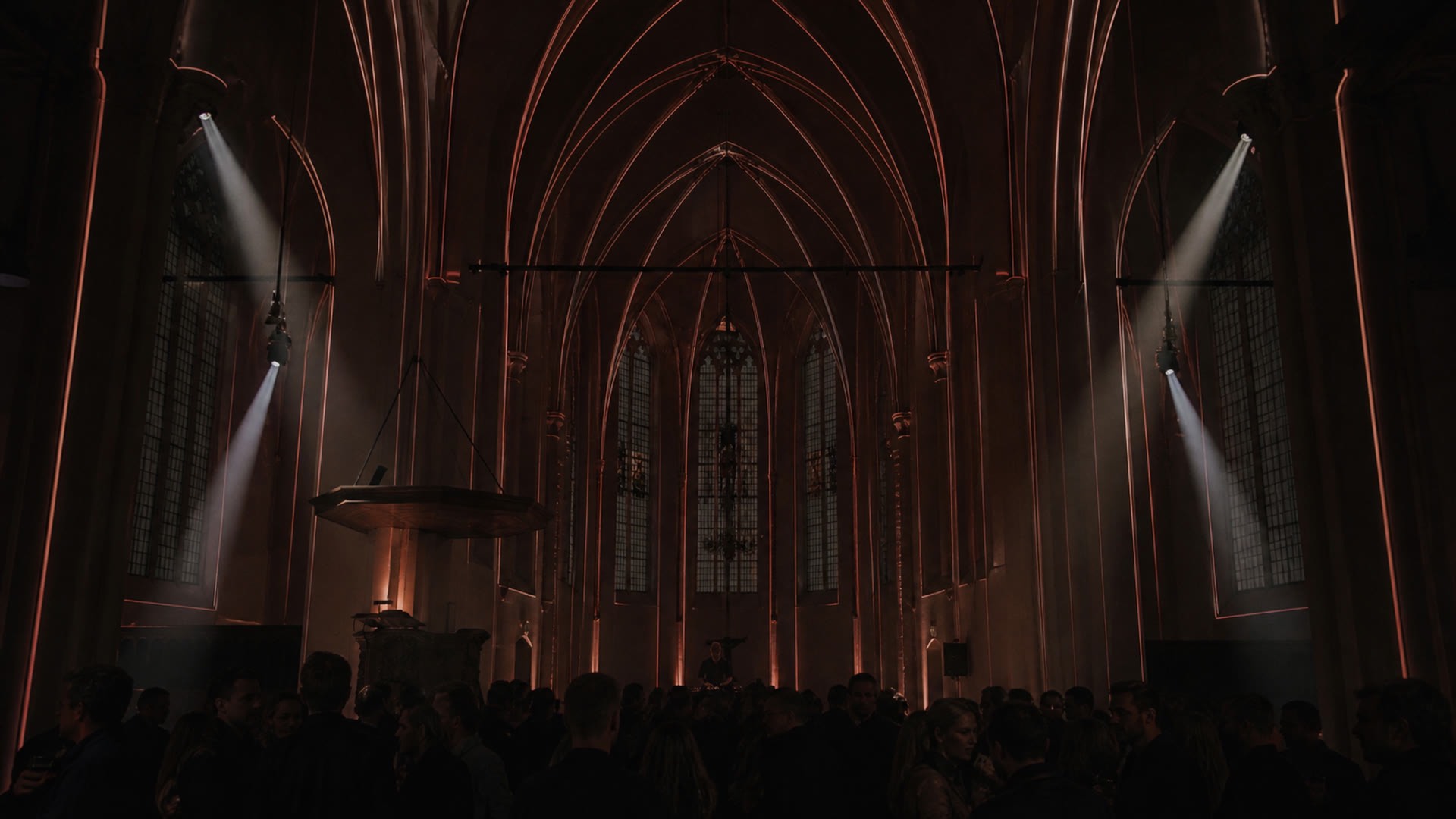 The architecture drawn in light. Piers and ribs traced in copper, beams cutting the haze, a crowd below in silhouette.