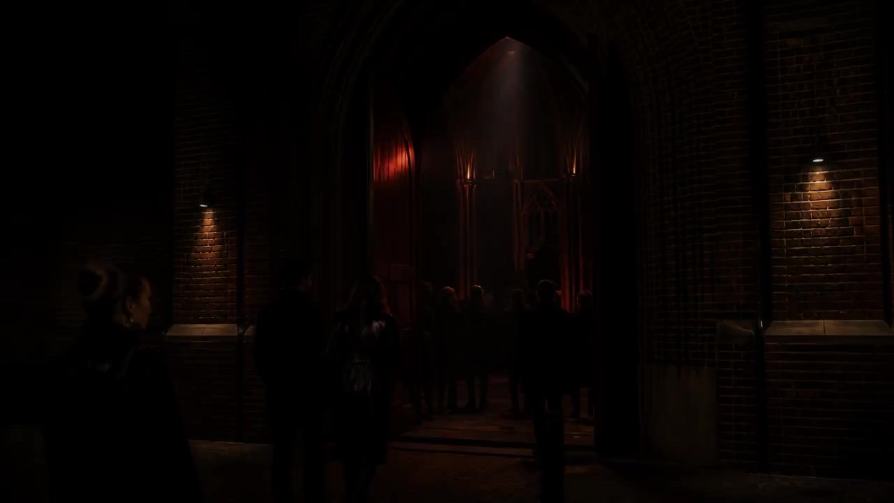 The lit arch of the church porch at night, guests in dark coats walking through it into a corridor of deep red light.