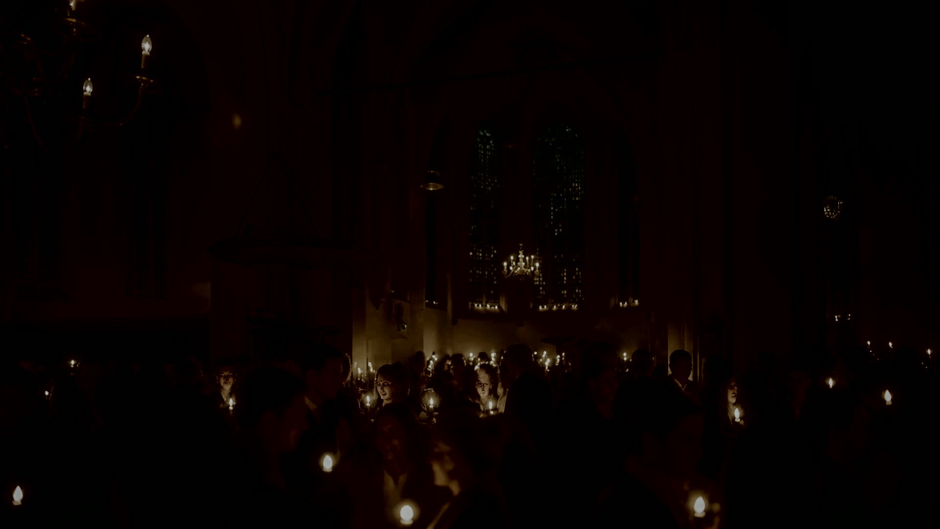 A crowd in near darkness carrying points of light, faces caught between them.